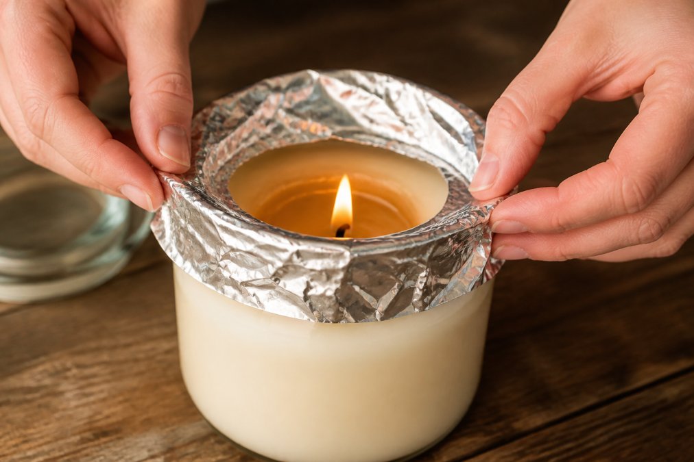 Hands carefully wrapping aluminum foil around the top rim of a tunneled candle jar, with the foil folded to direct heat downward while the wick remains visible and burning