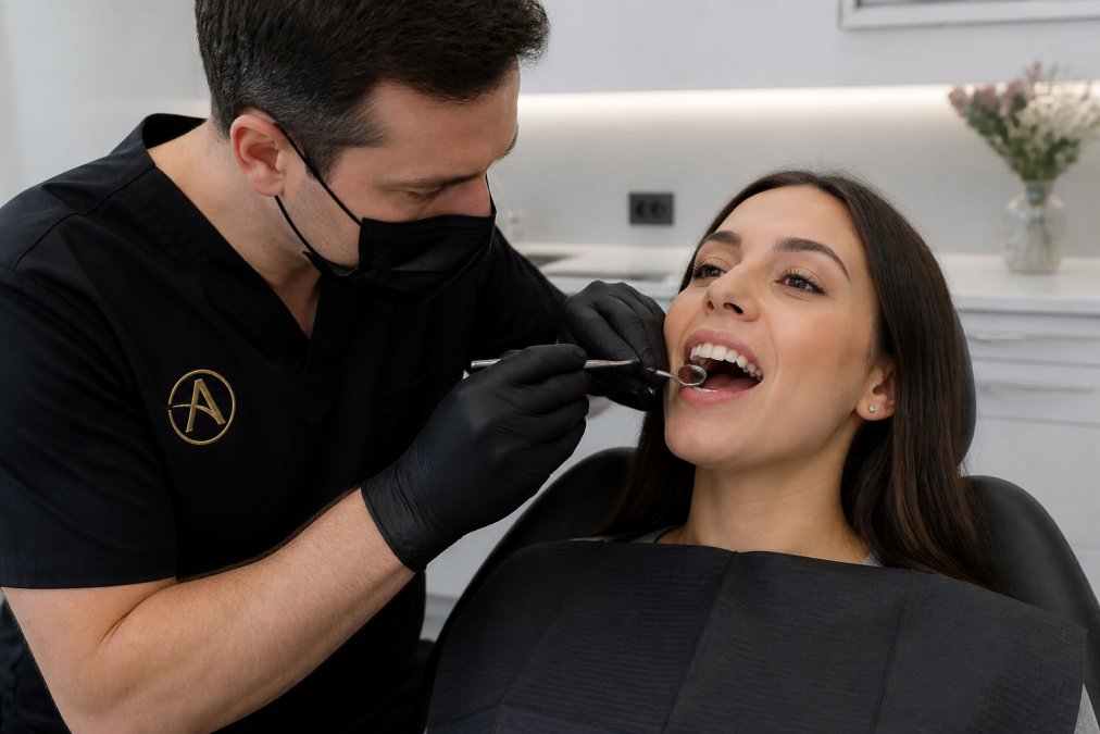 A dentist in gloves examining a patient's front teeth with a dental mirror in a bright, modern private treatment room, the patient reclined and calm