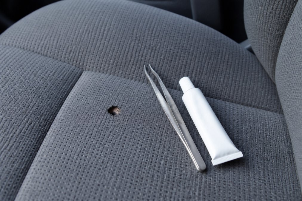 Close-up of a car seat with a small cigarette burn hole in grey fabric upholstery, with tweezers and a small tube of fabric adhesive resting on the seat beside it in natural daylight