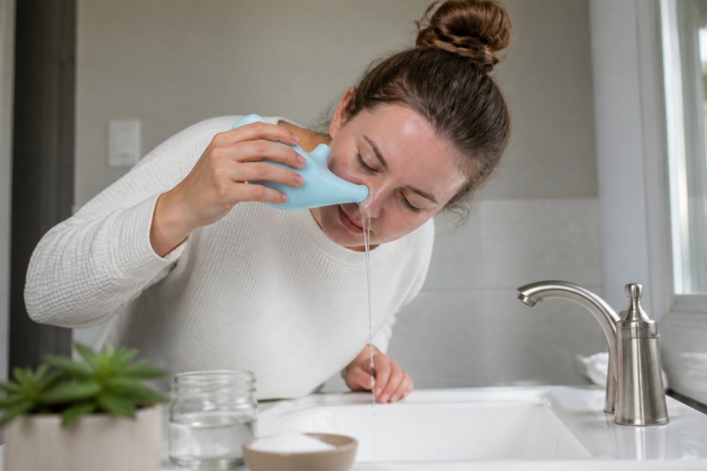 Person performing saline nasal irrigation at a sink using a neti pot, demonstrating proper head positioning and technique with natural bathroom lighting