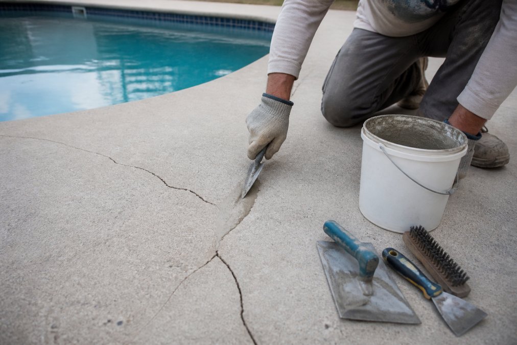 A worker in work gloves applying patching compound into a crack on a concrete pool deck using a trowel, with repair tools and a bucket of compound visible nearby on the deck surface