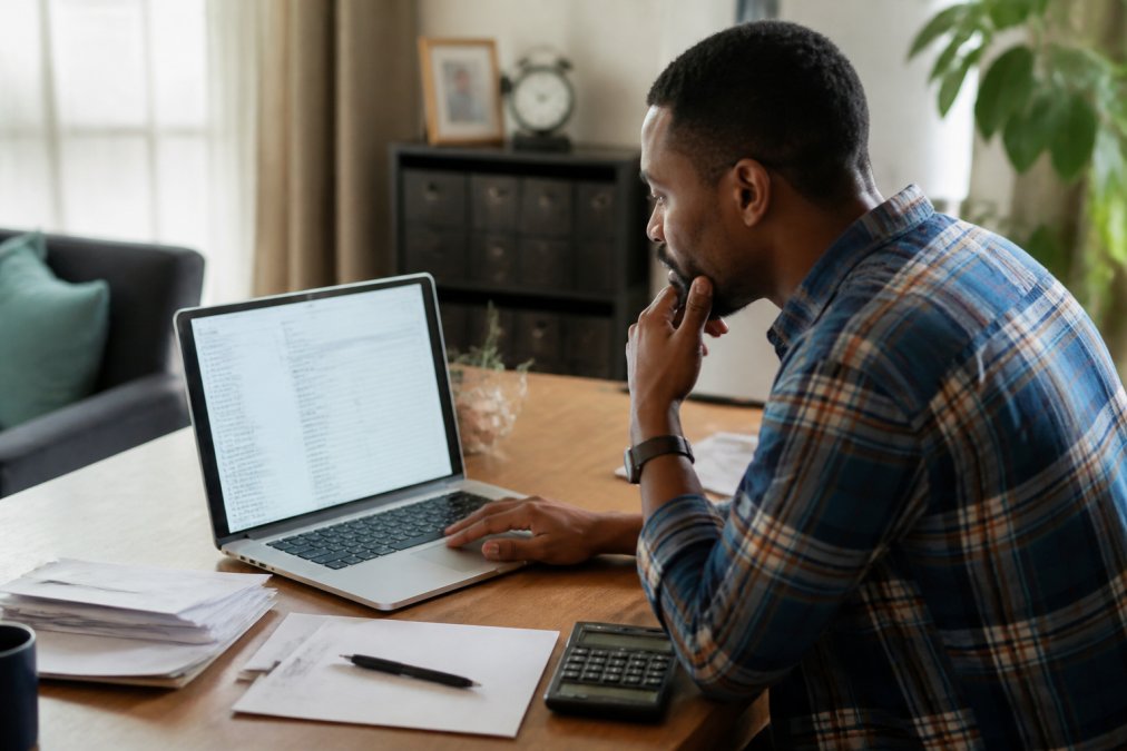 Person reviewing credit report on laptop at home desk with financial documents and pen nearby, studying account details with focused expression