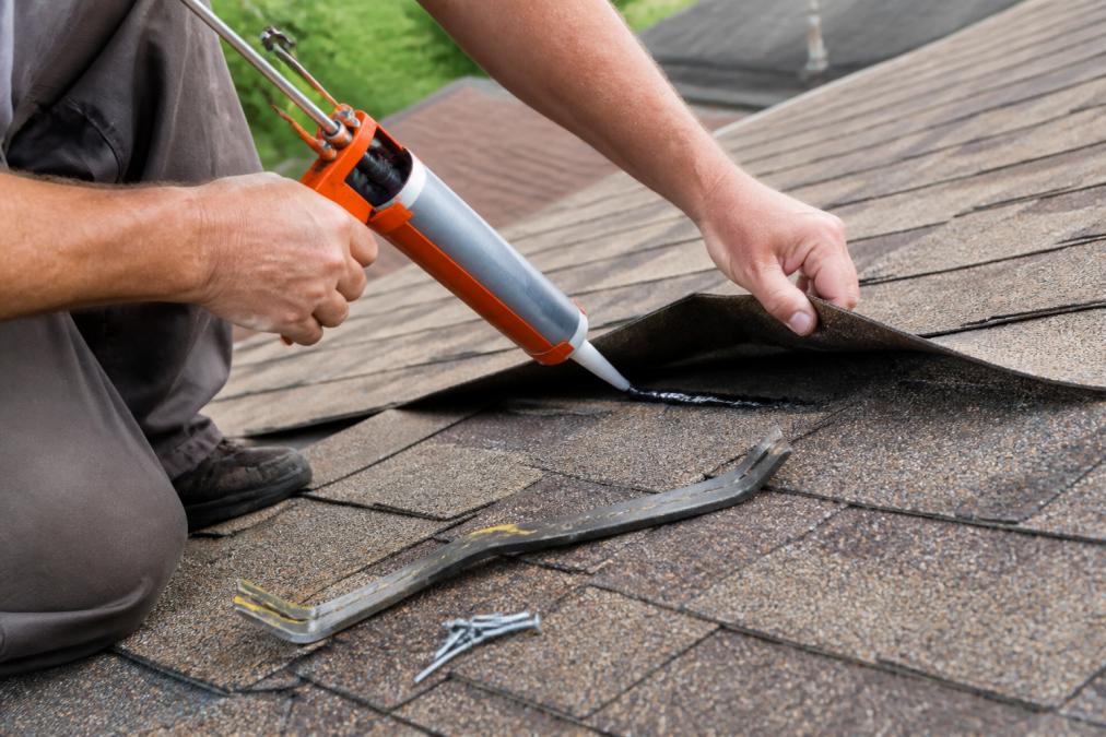 Close-up of a homeowner's hands using a caulking gun to apply roofing cement under a curled asphalt shingle tab on a residential roof, with a pry bar and roofing nails resting nearby on the shingles