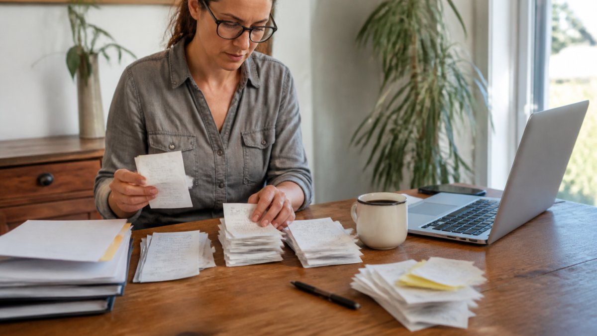 A small business owner at a wooden desk surrounded by organized stacks of receipts, bank statements, and a laptop showing a spreadsheet, with a coffee mug and pen nearby, natural window light