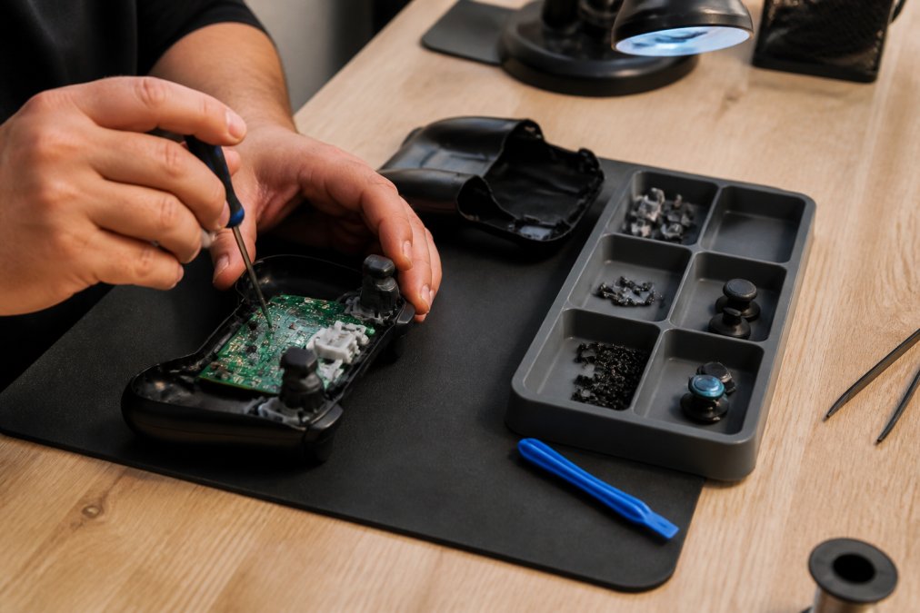 Close-up of hands using a precision screwdriver to open a game controller on a clean workbench, small screws and joystick parts arranged in a tray under bright task lighting