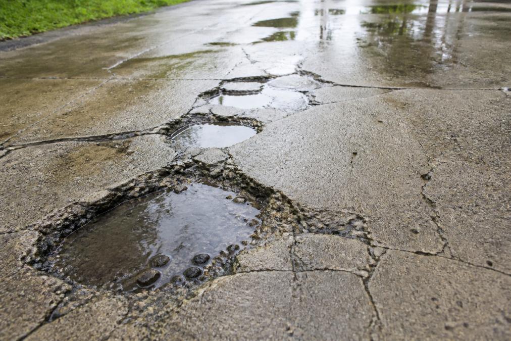 Close-up of a cracked and water-damaged concrete driveway with visible potholes, surface erosion, and standing water pooling in depressions after heavy rain, photographed under overcast natural light