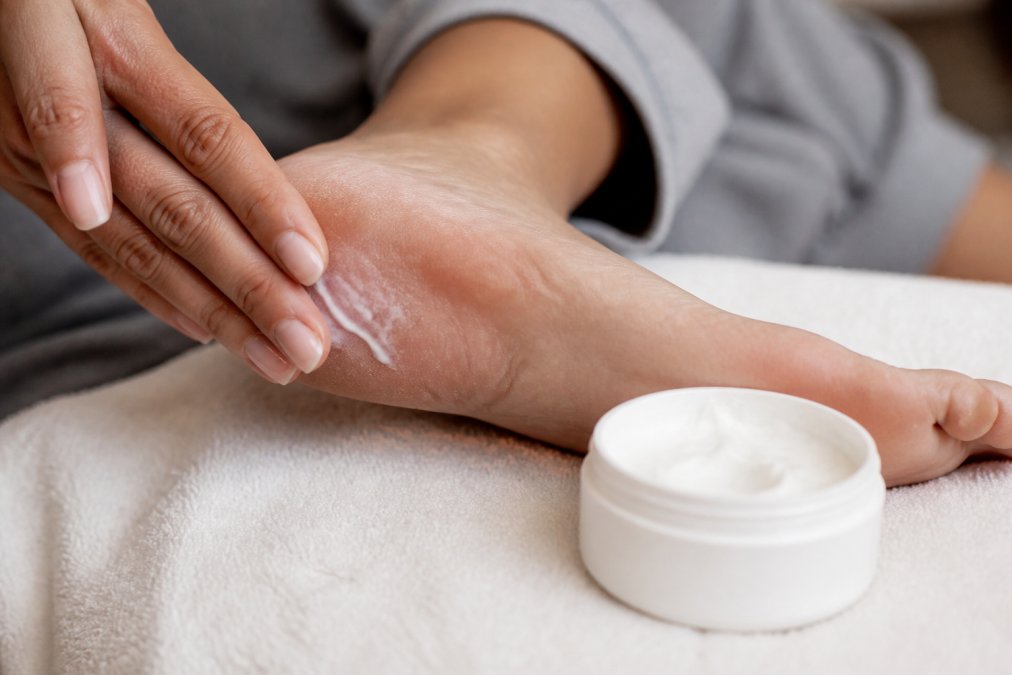 Close-up of hands applying a thick white foot cream to the heel of a foot, with an open jar of cream resting on a soft towel nearby, bathed in warm, soft lighting