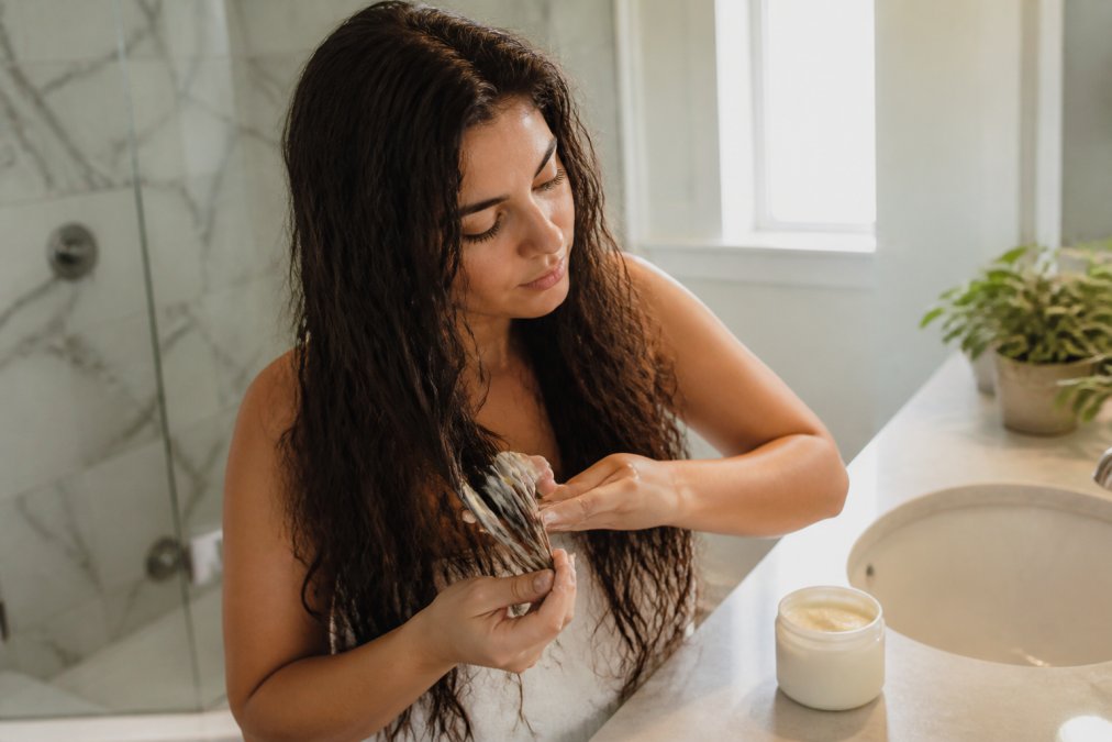 A woman with long, wavy hair applying a deep conditioning hair mask to her dry ends in a bright bathroom, focusing on the ends of her hair, with a jar of hair mask visible on the counter