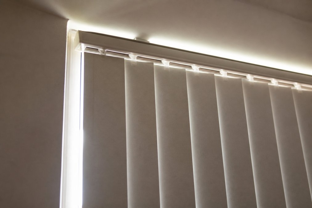 Close-up of a window with vertical blinds installed, showing visible light leakage around the side edges and gaps between the blind slats and window frame, demonstrating the light control problem homeowners face