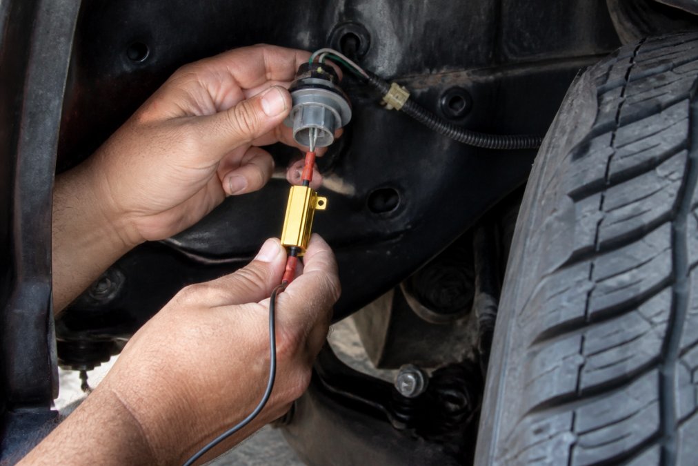 Close-up of hands wiring a load resistor to a vehicle's turn signal bulb socket near the wheel well, with visible wiring harness and connector clips under workshop lighting
