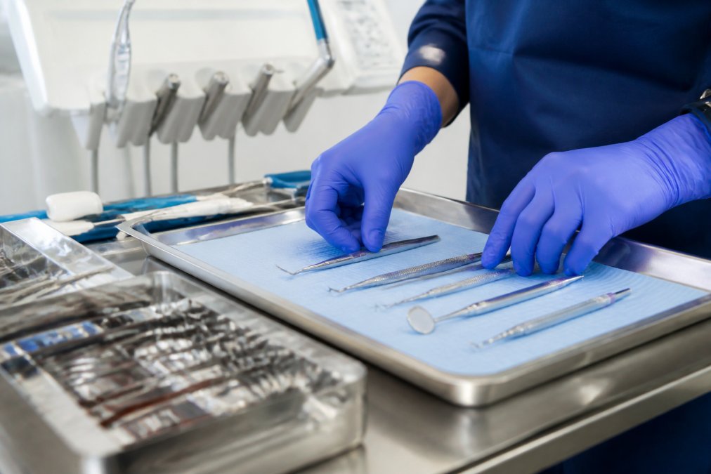 Close-up of a dental professional's gloved hands carefully arranging sterile instruments on a stainless steel tray in a clean, brightly lit treatment room, with organized surgical tools visible in the background Close-up of a dental professional's gloved hands carefully arranging sterile instruments on a stainless steel tray in a clean, brightly lit treatment room, with organized surgical tools visible in the background