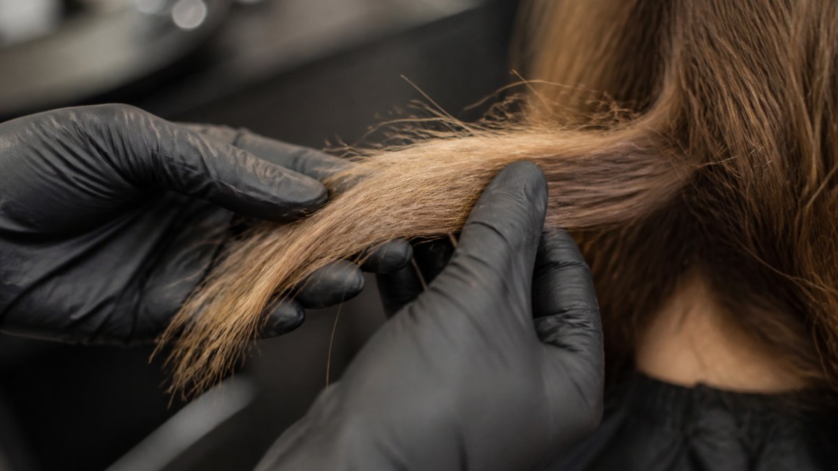 Close-up das mãos enluvadas de um cabeleireiro segurando uma mecha de cabelo seco e quebradiço com pontas duplas visíveis sob a luz brilhante do salão, mostrando textura áspera e quebra