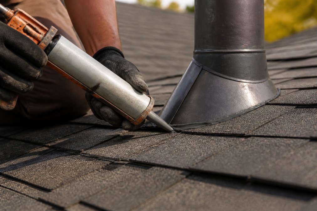 Professional roofer's hands applying roofing cement with a caulking gun to repair roof flashing on an asphalt shingle roof with afternoon sunlight