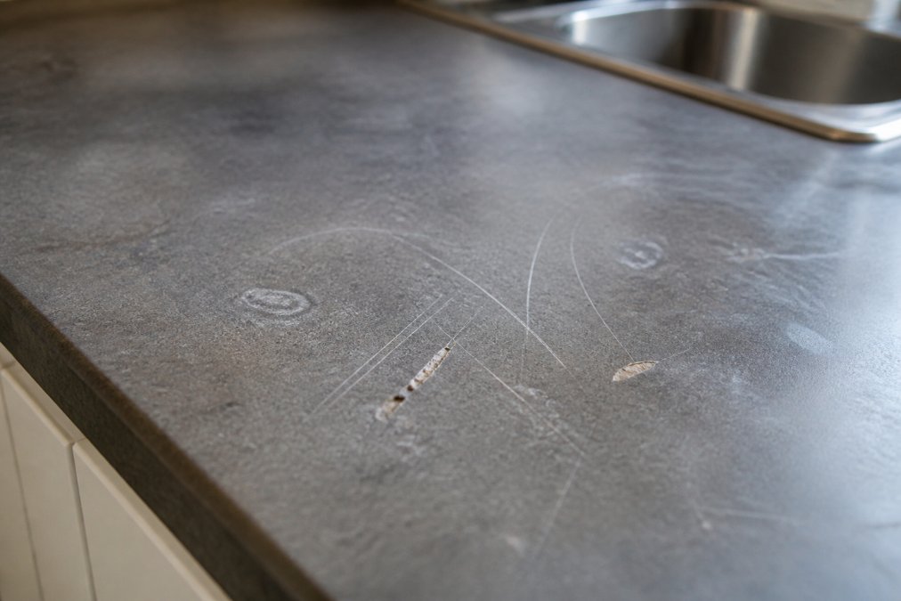 Close-up of a laminate worktop surface showing various scratch depths and damage types, with natural kitchen lighting highlighting the texture and finish