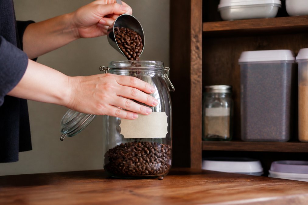 Hands carefully placing whole coffee beans into an airtight glass container with a handwritten date label visible, with a dark wooden pantry shelf and opaque storage containers in soft natural light