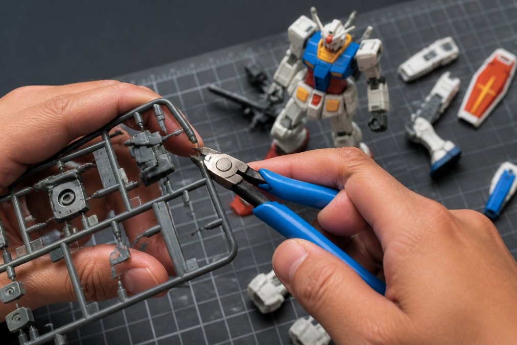Close-up of a builder's hands holding quality side cutters positioned at the correct 45-degree angle against a plastic runner, with finished Gundam parts visible in the background on a work mat