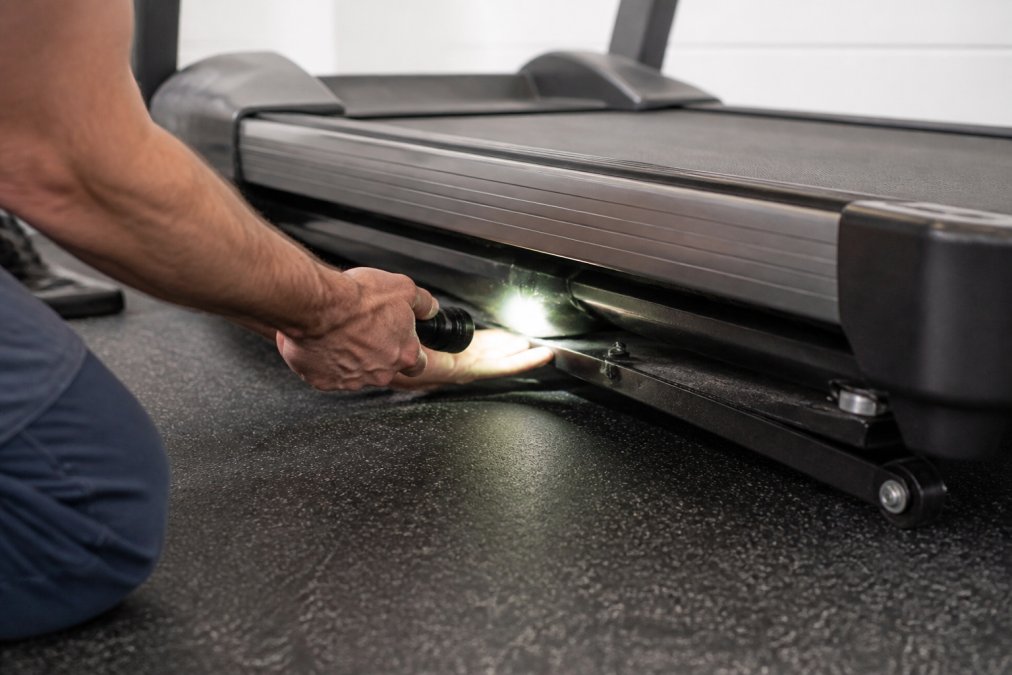 Close-up of hands using a flashlight to inspect the underside of a treadmill belt, checking for fraying, wear patterns, and debris accumulation on the running deck surface