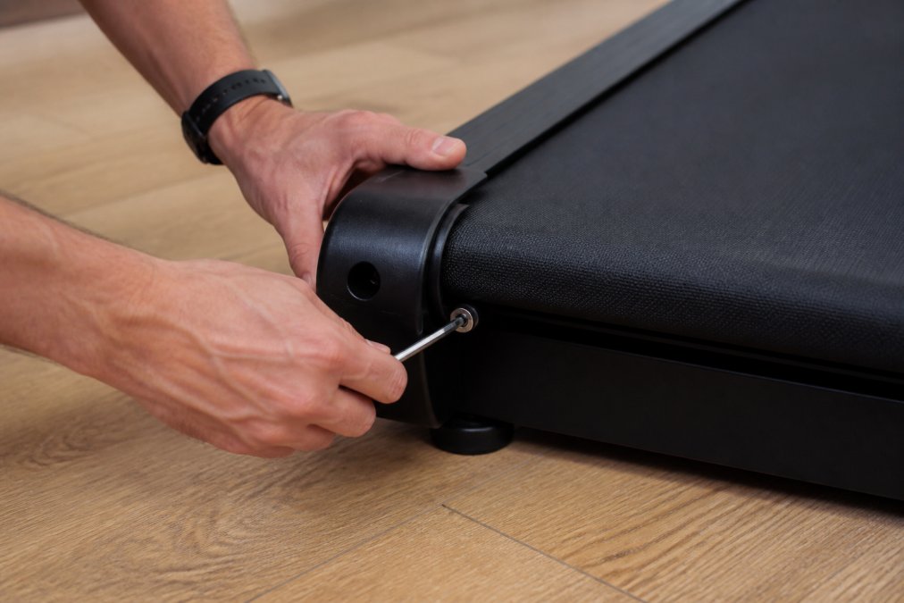 Person's hands using a hex wrench to turn the tension bolt at the rear corner of a treadmill, demonstrating proper hand position and counterclockwise rotation for loosening