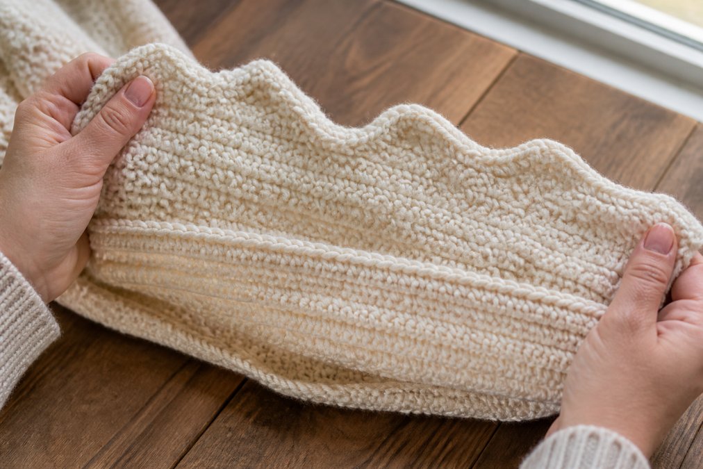 Close-up of hands holding a crochet blanket with visibly wavy and uneven edges, showing the contrast between a straight section and a curved section, soft natural window lighting on a wooden table