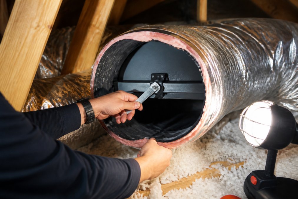 Professional HVAC technician's hands adjusting a metal air damper lever inside flexible ductwork in an attic space, with insulation visible in background and bright work light illuminating the adjustment
