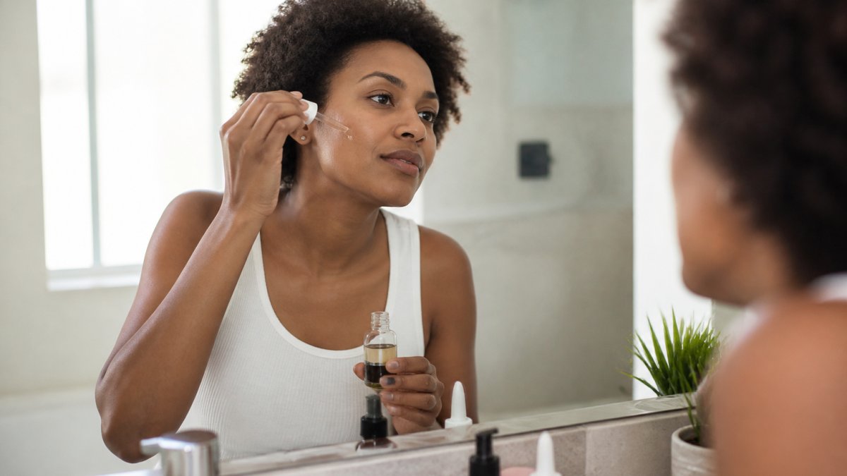 A woman with melanin-rich skin applying a treatment serum to her face in a bright bathroom, looking into a mirror with skincare products on the counter