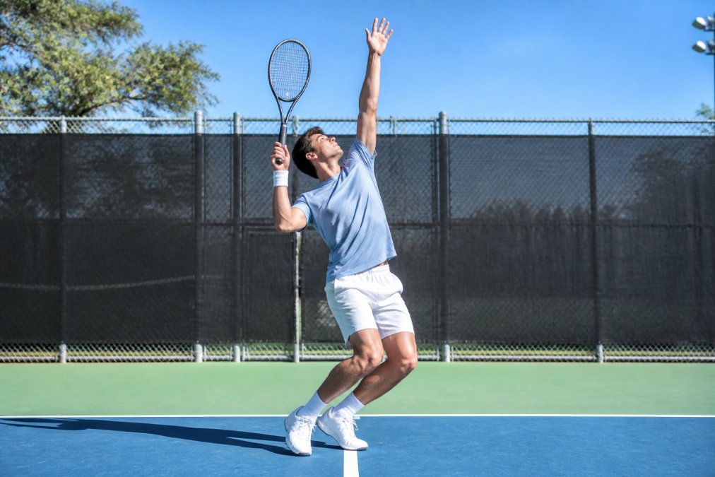 A tennis player at the trophy position on an outdoor hard court, racket arm fully extended upward, knees bent, body coiled sideways, captured mid-serve motion in bright daylight