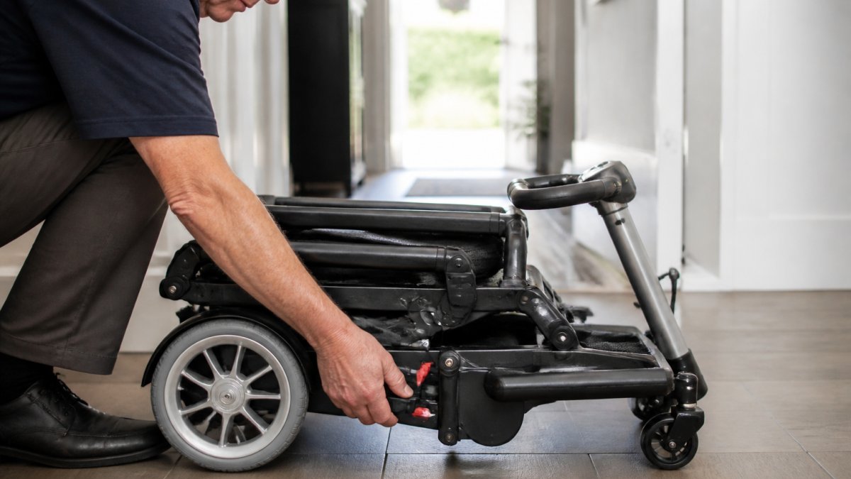 Close-up of a person's hands pressing the release lever on a folded lightweight folding mobility scooter in a hallway, with the seat backrest already lowered and the tiller tilted forward