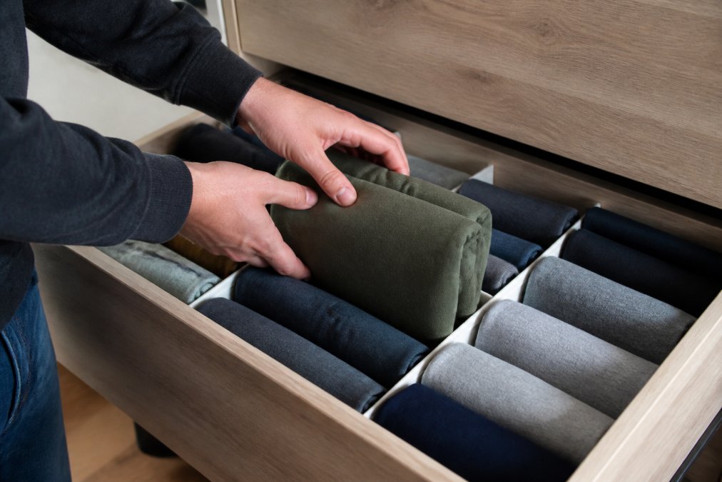 A person's hands placing a folded hoodie upright into a dresser drawer with dividers, the front edge of the hoodie visible like a file folder, surrounded by other neatly filed hoodies in a tidy drawer with soft natural light