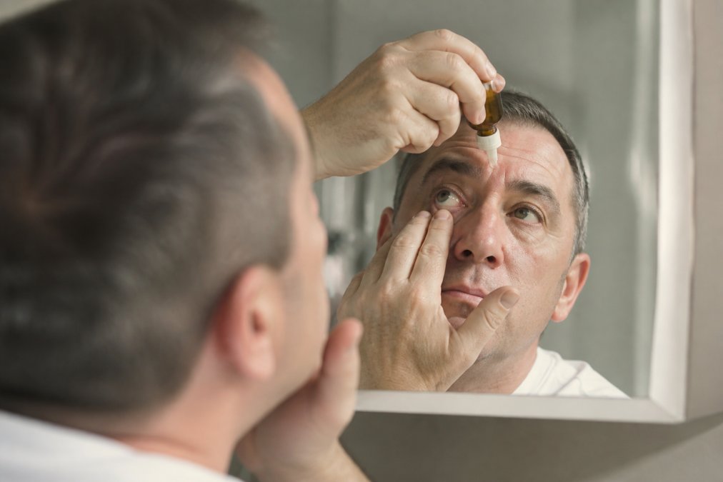 Person carefully using prescription drops for eye surgery aftercare while looking into a bathroom mirror