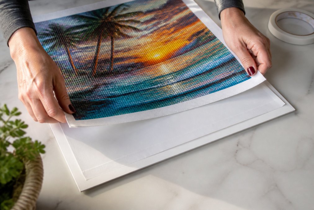 Close-up of hands centering a finished diamond painting canvas onto a white backing board, using double-sided tape on the board edges, bright natural light on a clean craft table
