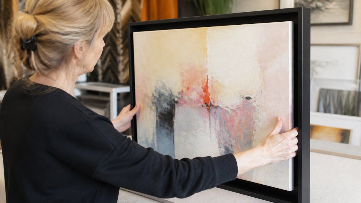 Professional demonstrating careful positioning of stretched abstract canvas into floater frame, showing proper alignment and even spacing on all sides with hands gently guiding canvas into place