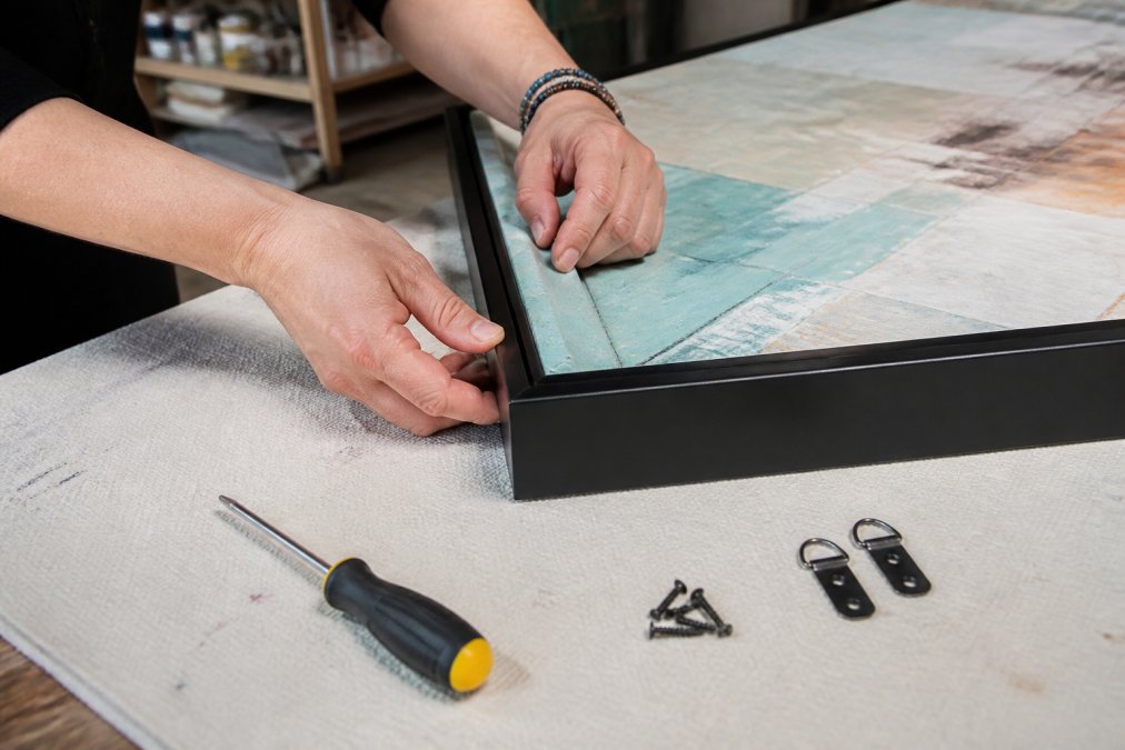 Close-up of hands attaching a slim black floater frame to a stretched abstract canvas on a clean workbench, with a screwdriver and D-rings visible nearby