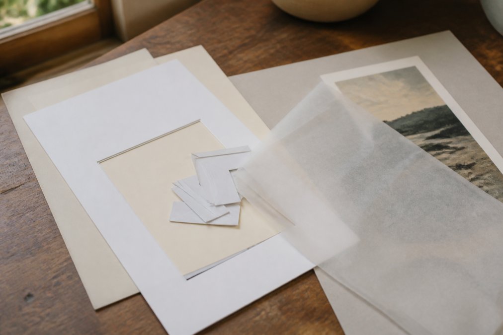 Close-up of framing materials laid out on a wooden worktable: acid-free mat board in cream and white, pH-neutral mounting corners, a translucent dust cover sheet, and a sheet of archival backing board with a fine art print partially visible underneath, in warm natural window light