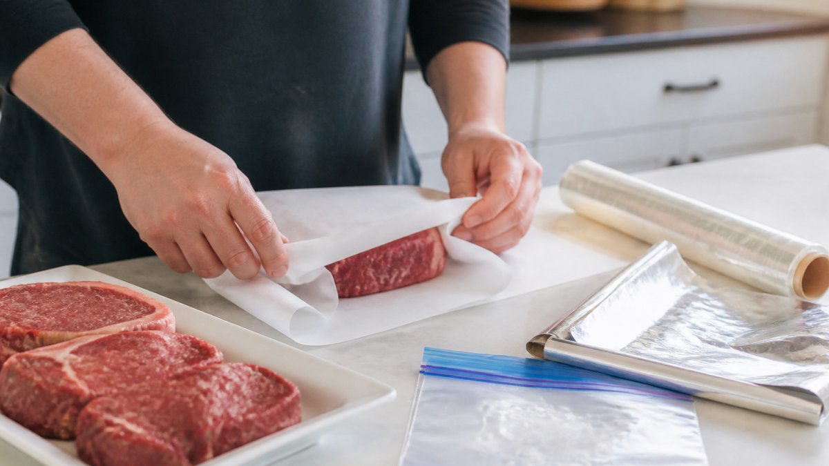 Hands wrapping a cut of raw beef tightly in freezer paper on a clean kitchen counter, with plastic wrap, freezer bags, and a roll of foil nearby