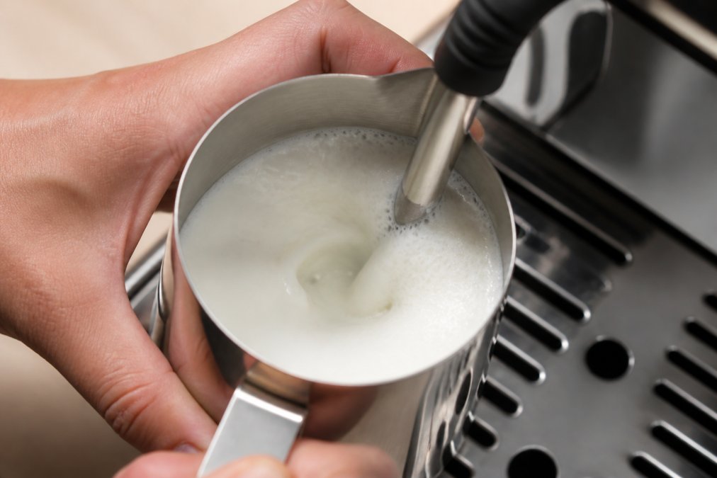Close-up of a barista's hand holding a stainless steel pitcher at an angle under the steam wand of an espresso machine, milk creating a visible vortex with fine bubbles, bright kitchen lighting