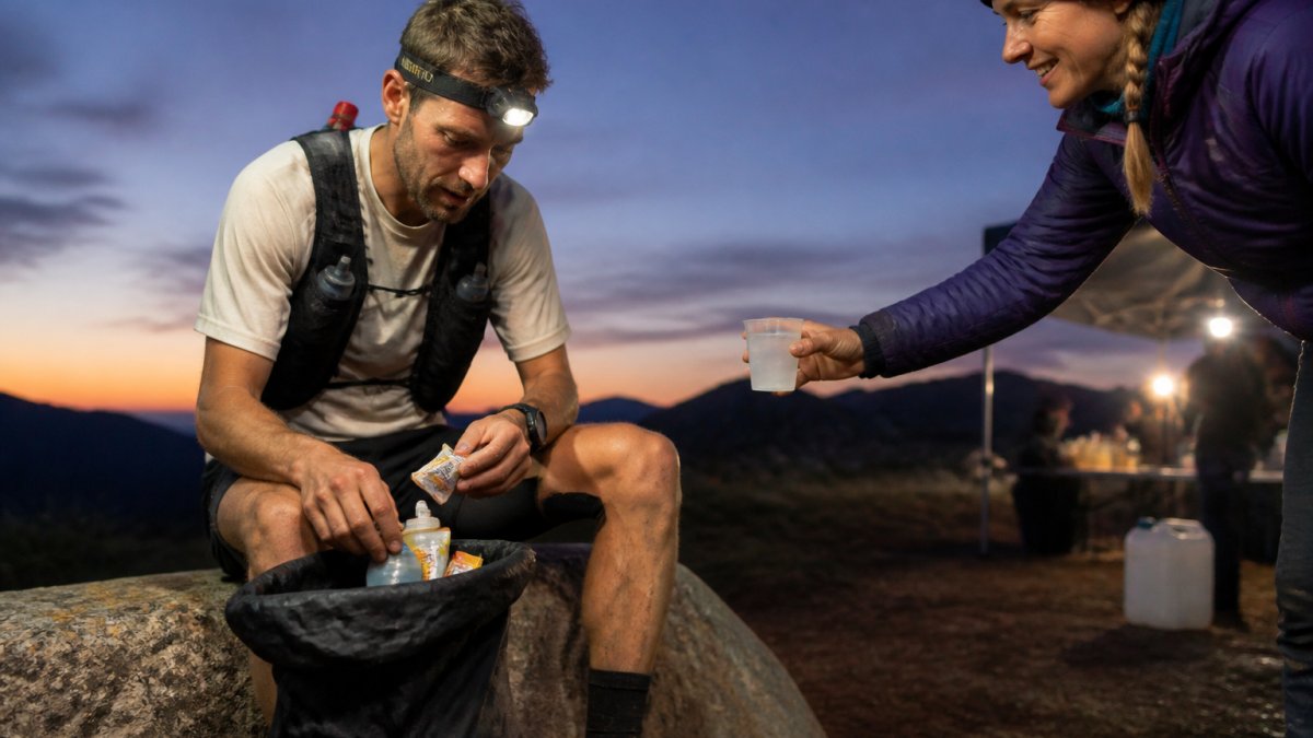Trail runner sorting energy gels and drink bottles at a dusk aid station while a volunteer hands over water