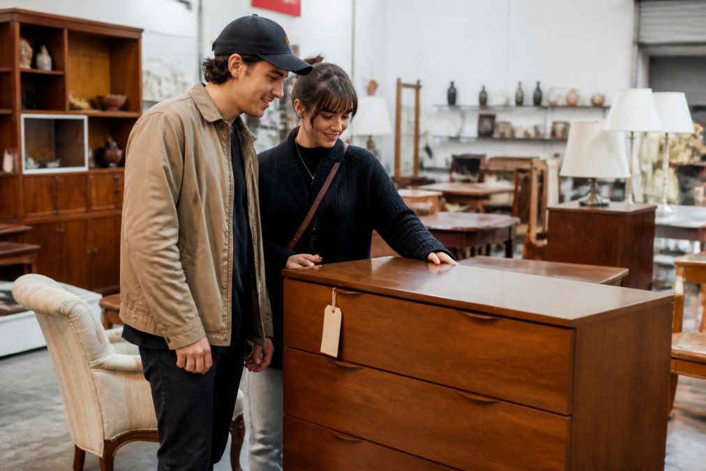 A young couple shops for a mid-century dresser at a thrift store to furnish their apartments on budget.