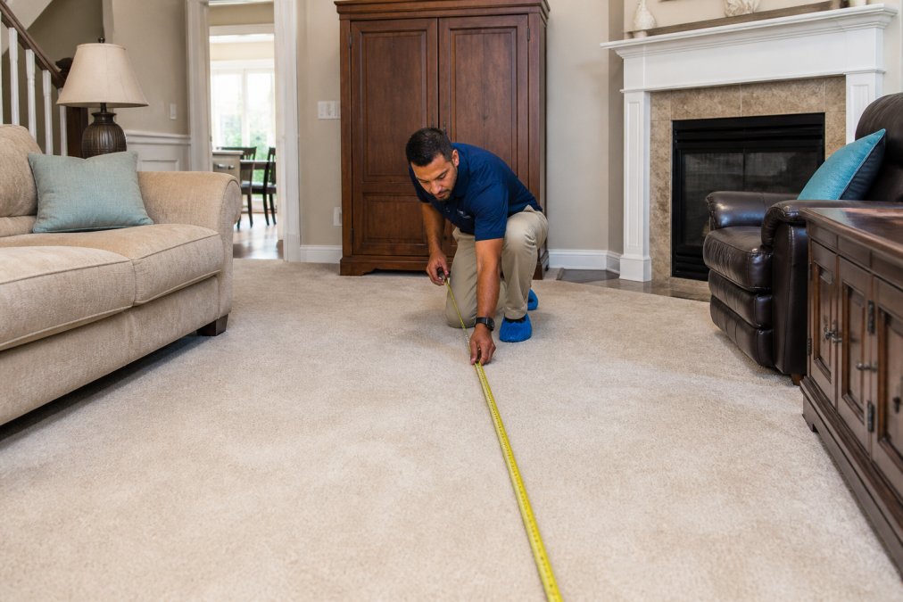 Professional carpet cleaner measuring room dimensions with tape measure in well-lit residential living room with furniture moved to sides