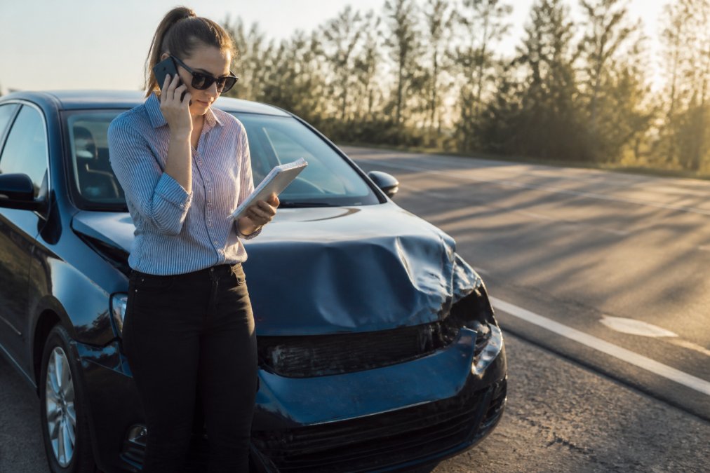 A person standing beside a damaged car on the roadside, speaking calmly on a mobile phone with a notepad in hand, afternoon light casting long shadows across the road