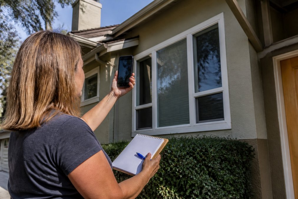 Homeowner photographing their home's exterior windows with a smartphone, documenting window condition and accessibility while holding a notepad with measurements for a quote request