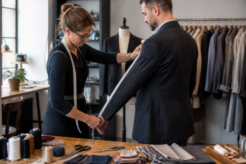 A seamstress measuring a customer's jacket sleeve in a bright tailoring studio, with fabric swatches and sewing tools visible on a wooden table