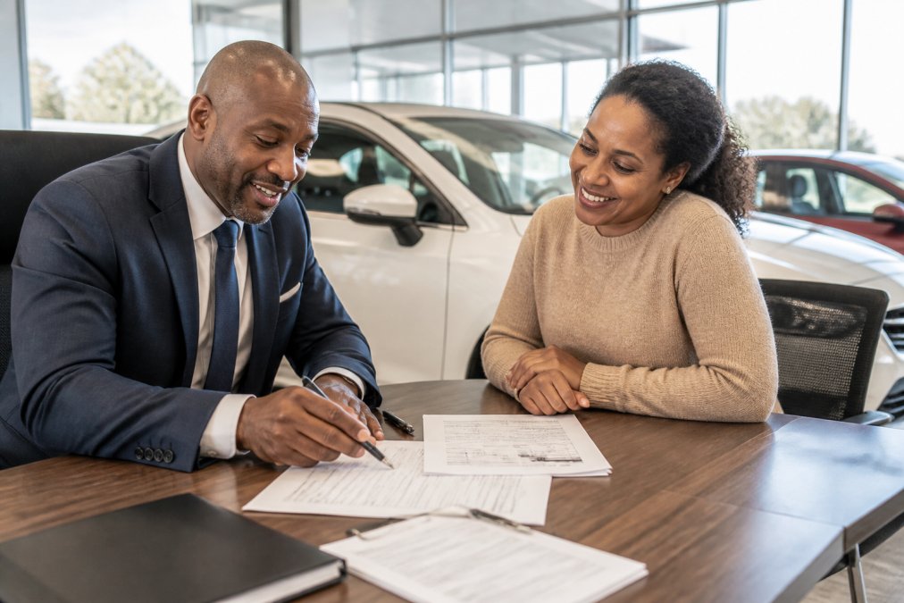 Professional sitting at desk reviewing loan application documents with customer in bright, welcoming dealership office