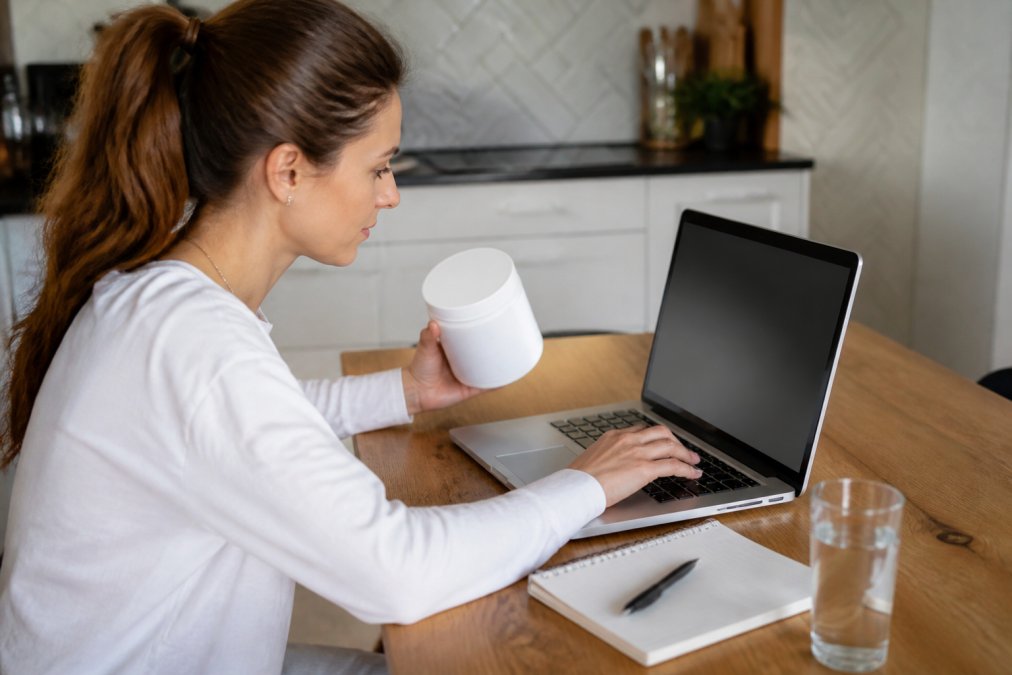 A person sitting at a kitchen table with a laptop, typing a refund request email while holding a collagen supplement container nearby, with a notepad and pen on the table