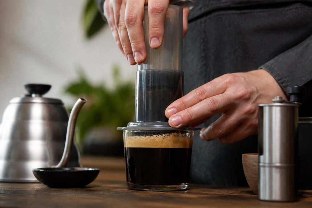 Hands pressing a coffee maker onto a glass cup to reveal a thick layer of golden-brown crema on top of the brewed coffee