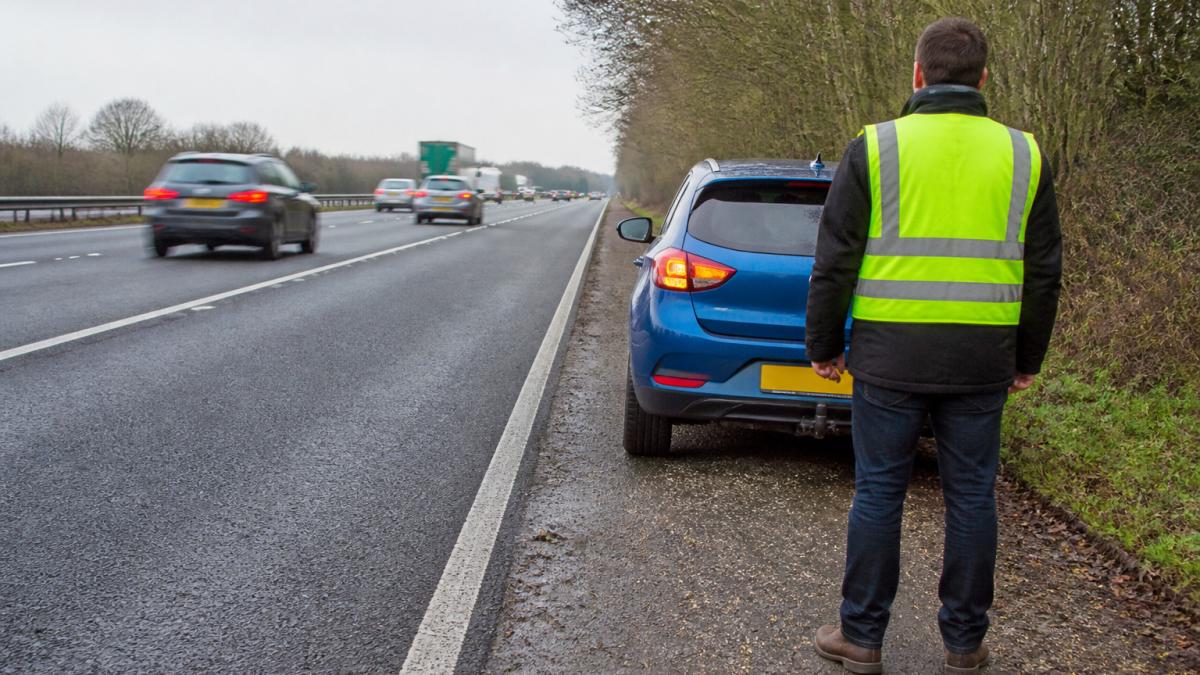 Driver in a high-visibility vest standing safely behind a car on a motorway hard shoulder awaiting vehicle recovery
