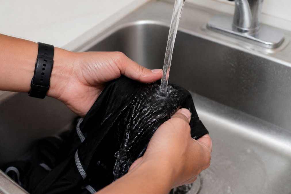 Close-up of hands holding moisture-wicking athletic gear under running water during a rinse cycle, showing water flowing through the fabric