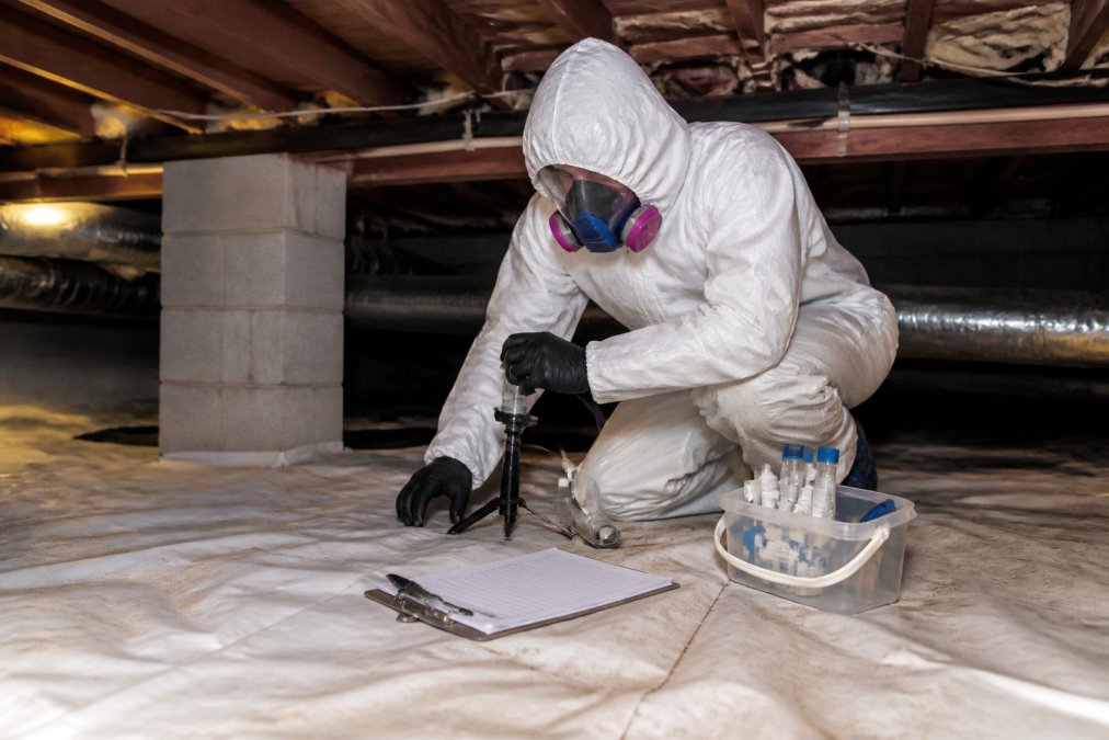 Professional mold inspector in protective white suit and respirator collecting air samples with specialized equipment in a residential crawlspace, with test containers and documentation clipboard visible on the floor beside them