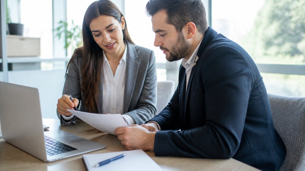 A homebuyer sitting across a desk from a loan officer in a bright office, reviewing paperwork together with a laptop open and a notepad of questions