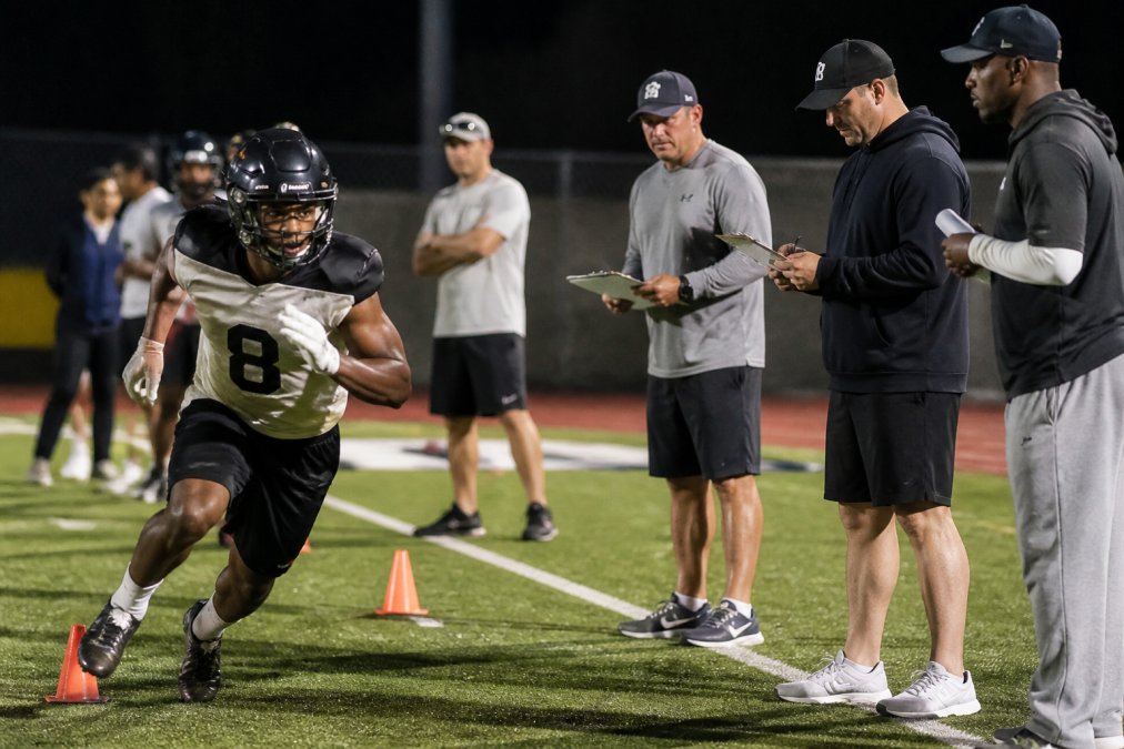 A high school football player in full pads running a drill at a college showcase camp, with coaches holding clipboards and evaluating on the sideline under bright stadium lights