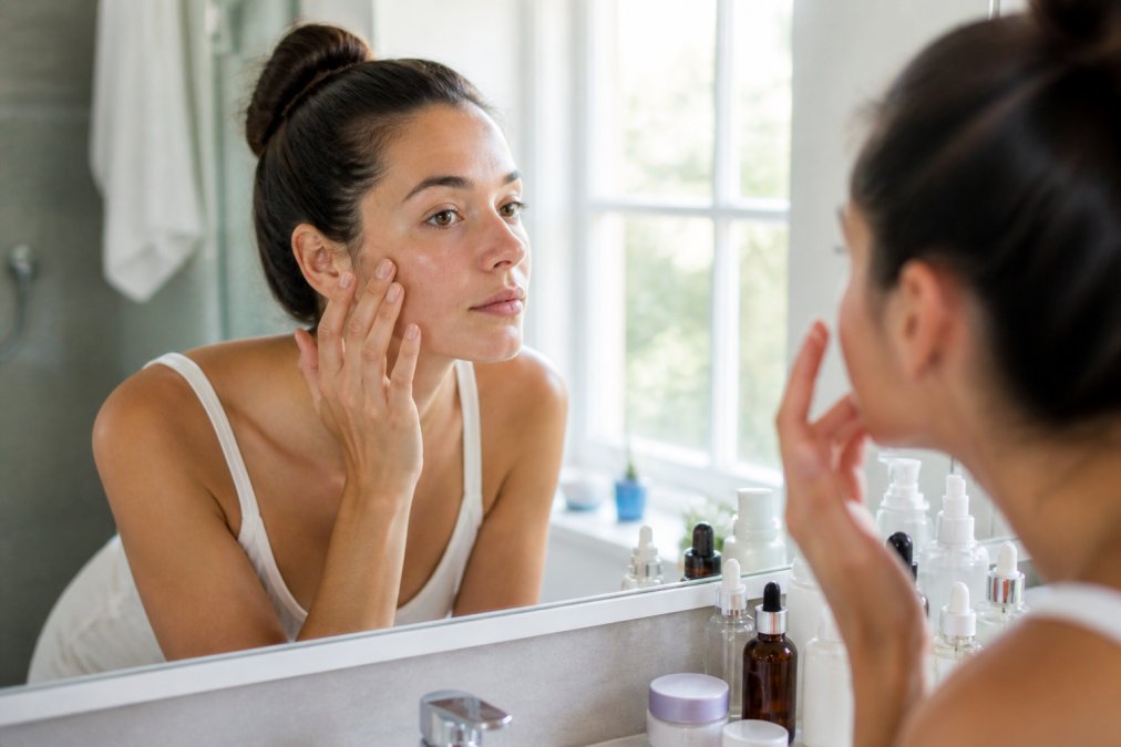 A woman in her late 20s examining her skin in a bathroom mirror with natural light, skincare products arranged neatly on the counter beside her